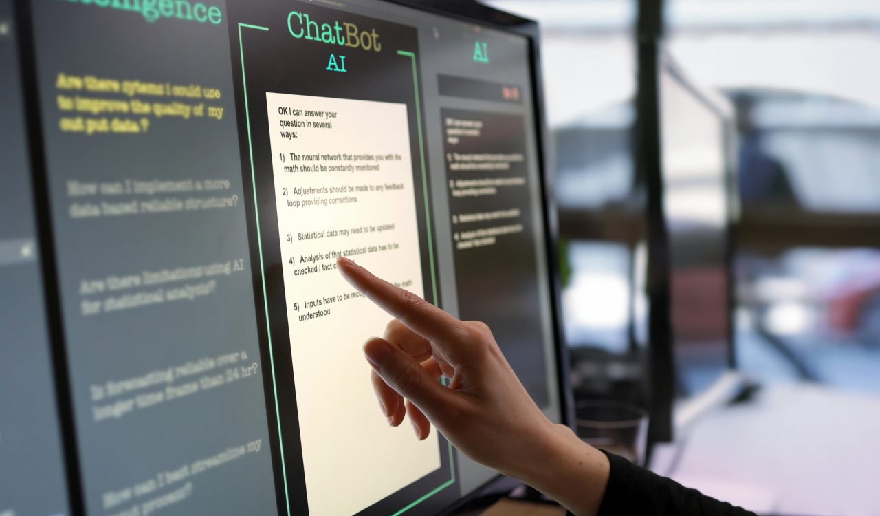 A touchscreen monitor being used in an open plan office. A woman’s hand is asking an artificial intelligence chatbot pre-typed questions and the artificial intelligence website is answering.