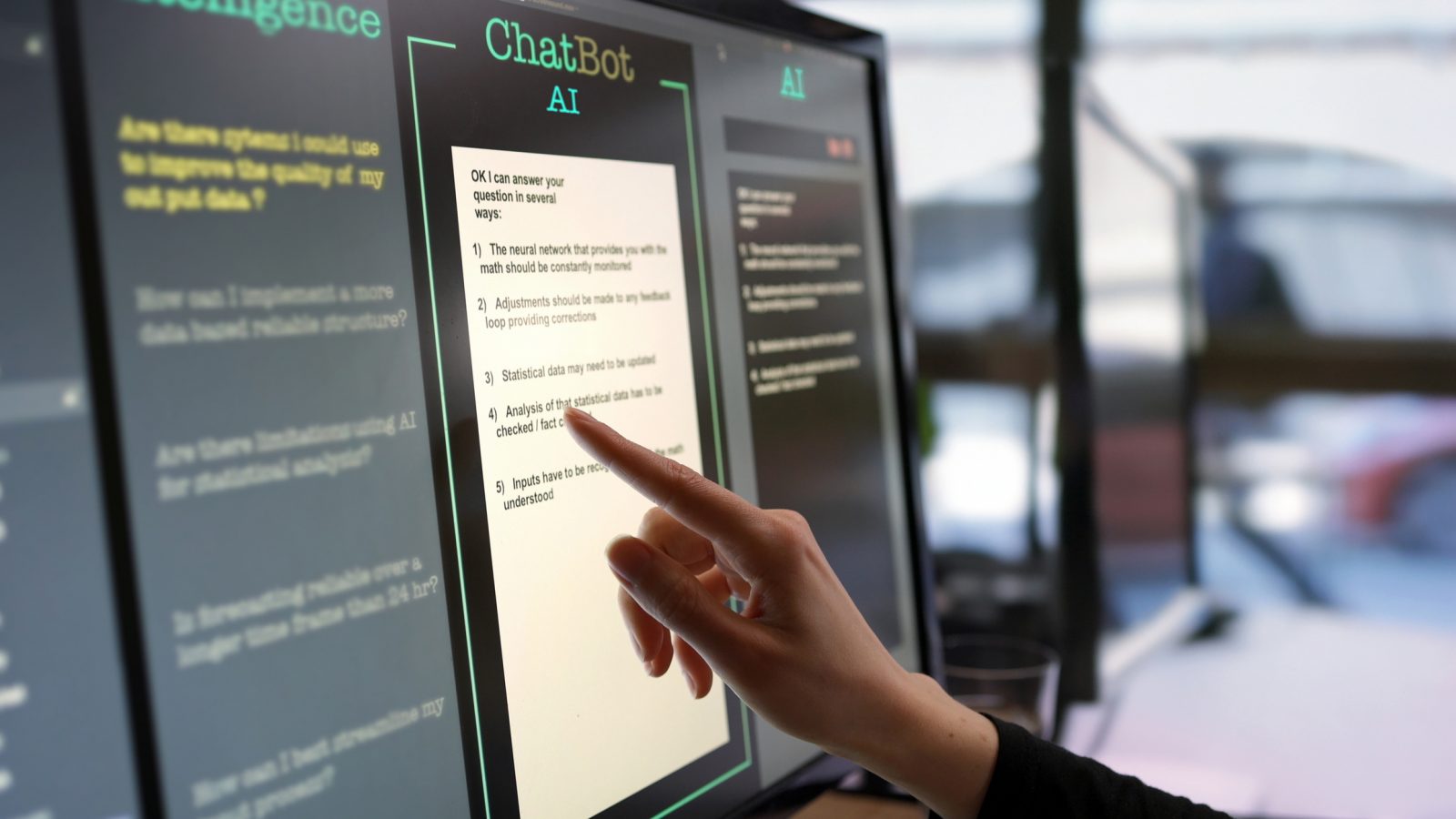 A touchscreen monitor being used in an open plan office. A woman’s hand is asking an artificial intelligence chatbot pre-typed questions and the artificial intelligence website is answering.