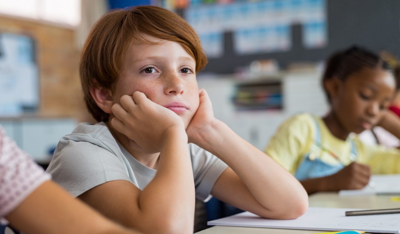 A boy sits with his head resting on his hands, with his elbows on a desk.