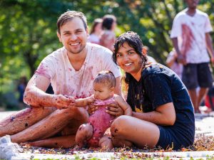 A young family poses for a photo while covered in grapes while seated on the ground outdoors.
