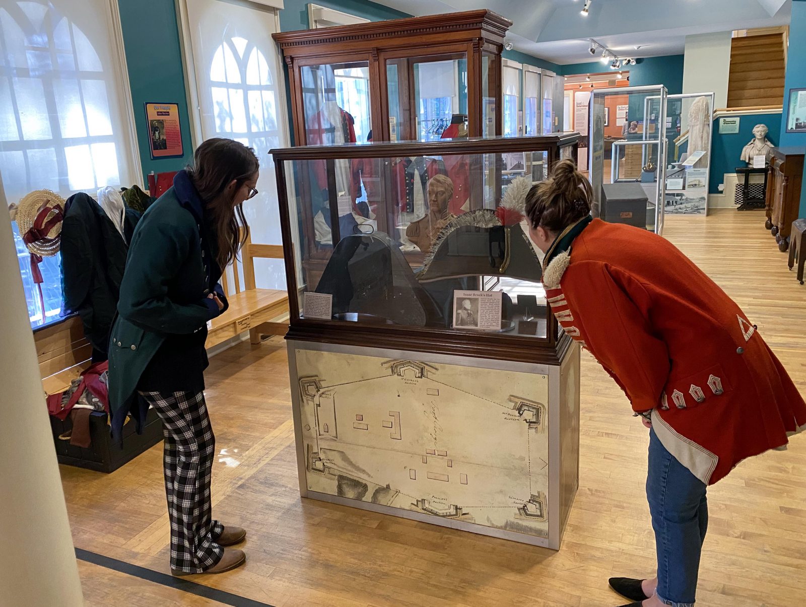 Two women with their backs to the camera are bending over to read signs in a glass display case featuring artifacts from the War of 1812, with other display cases in the background.