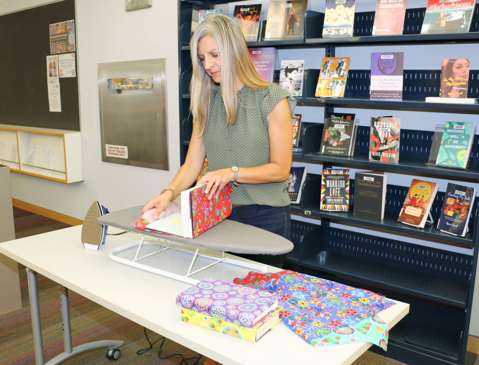 A woman places books within decorative cloth wraps while standing at a table in an indoor library.