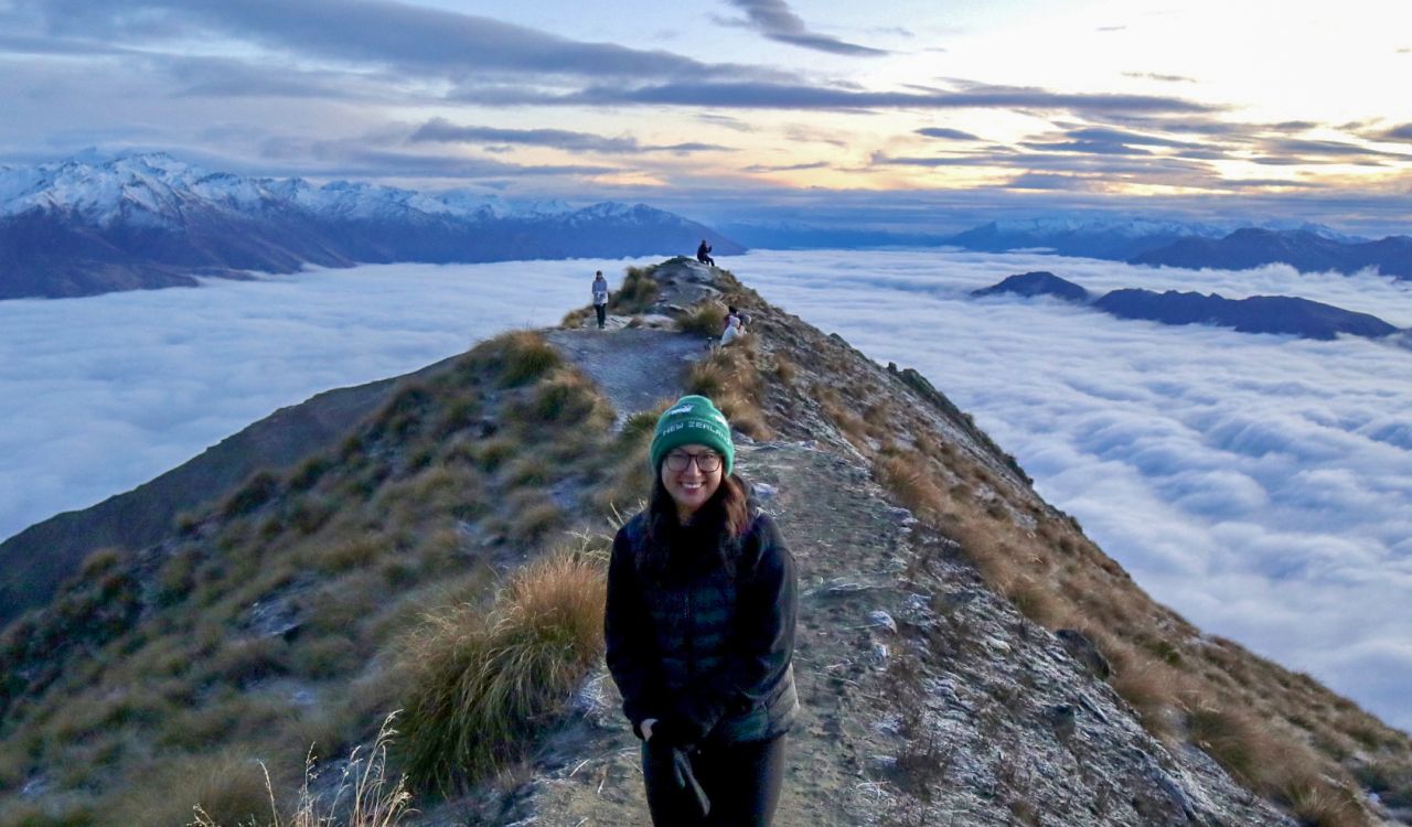 Brock University graduate student Jessica Liem stands on the summit of a mountain in Wanaka, New Zealand called Roy’s Peak. She is wearing warm clothing, including a toque, sweater and gloves. Clouds envelop the mountain, as the sun rises over Lake Wanaka.