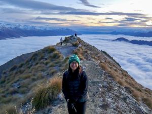 Brock University graduate student Jessica Liem stands on the summit of a mountain in Wanaka, New Zealand called Roy’s Peak. She is wearing warm clothing, including a toque, sweater and gloves. Clouds envelop the mountain, as the sun rises over Lake Wanaka.