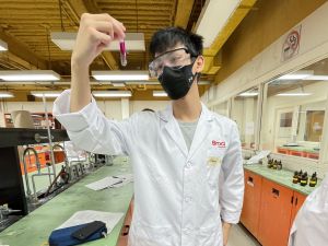 Former Brock University student Liam Jacob Zisman is in a Brock University science lab in the Fall of 2021. He is wearing a white Brock University lab coat, safety glasses and a face mask while looking at a test tube of clear liquid mixing with a red liquid.