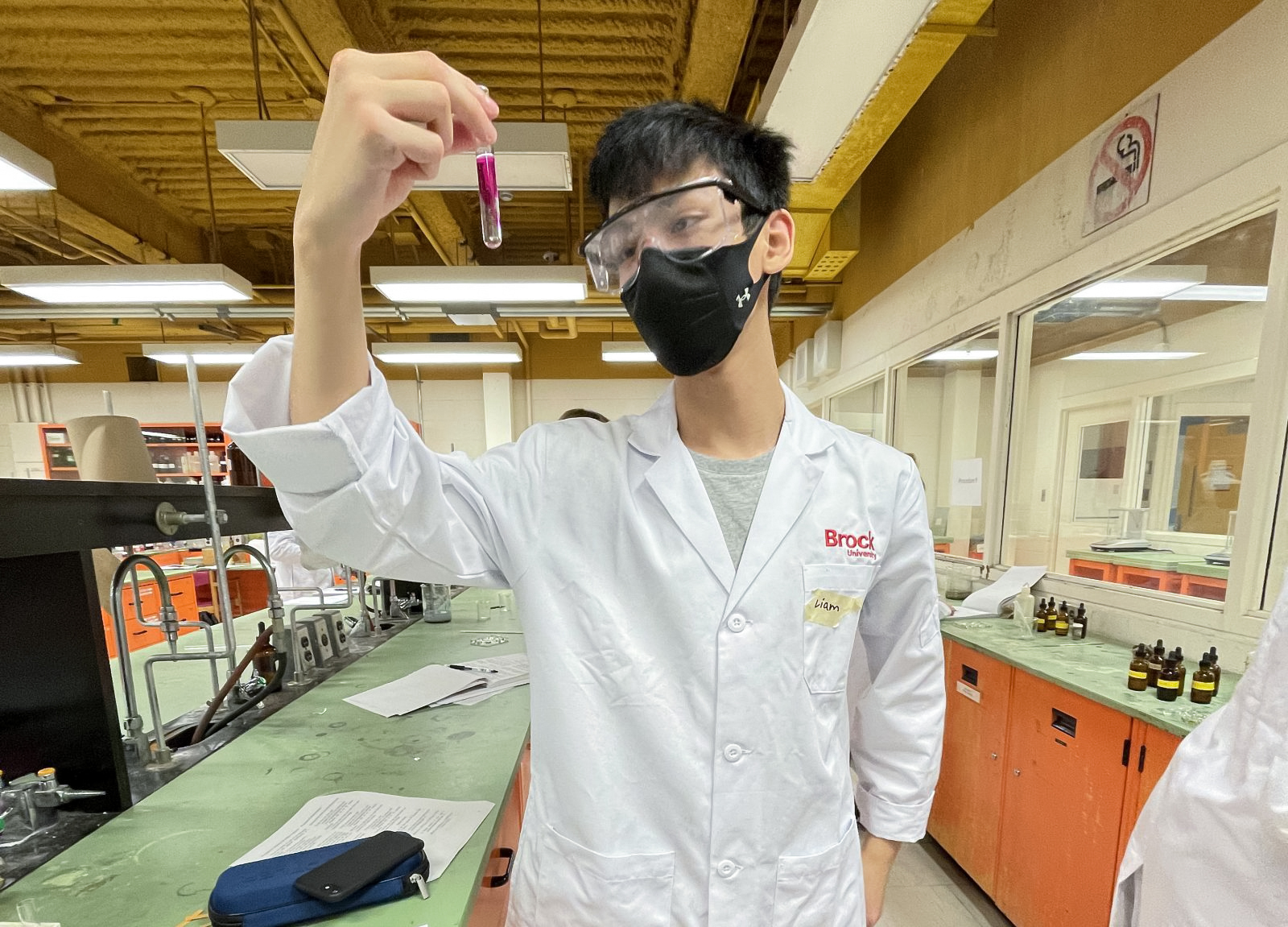 Former Brock University student Liam Jacob Zisman is in a Brock University science lab in the Fall of 2021. He is wearing a white Brock University lab coat, safety glasses and a face mask while looking at a test tube of clear liquid mixing with a red liquid.