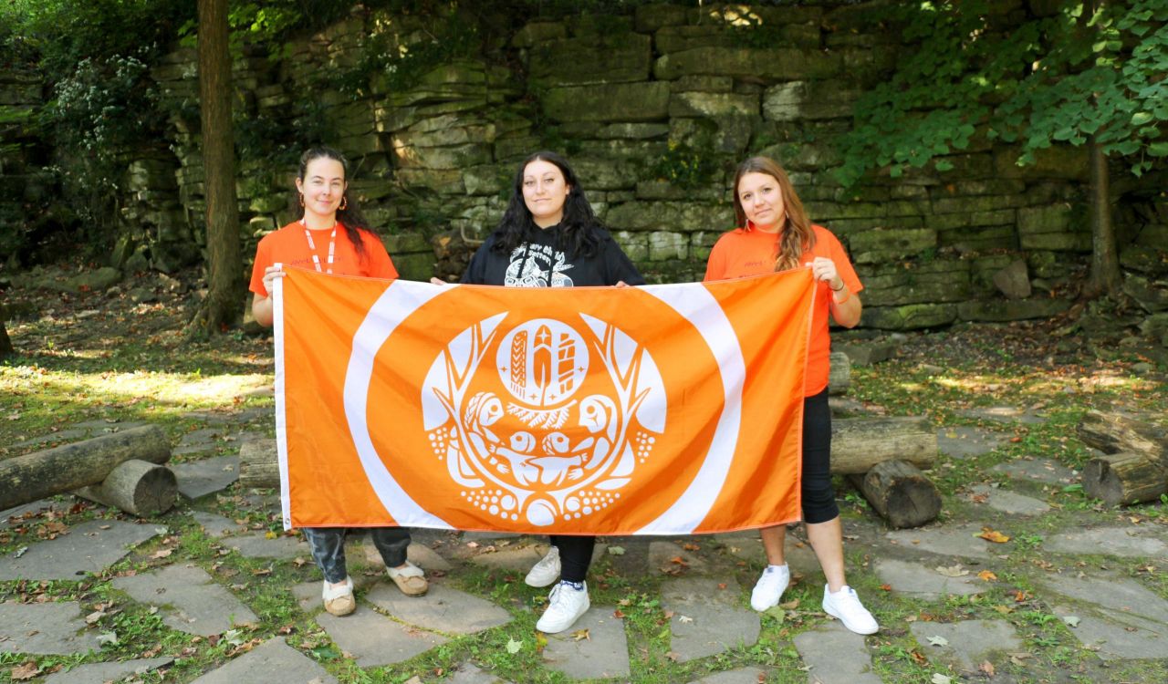 Two women in orange shirts stand on either side of a woman in a black shirt, while all three hold an orange flag in front of an outdoor wall made of rocks. 
