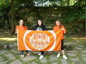 Two women in orange shirts stand on either side of a woman in a black shirt, while all three hold an orange flag in front of an outdoor wall made of rocks.