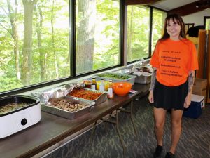 A woman in an orange shirt stands next to a table full of food.