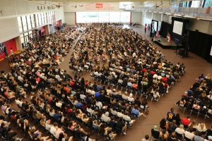 An indoor gymnasium full of thousands of seated university students.