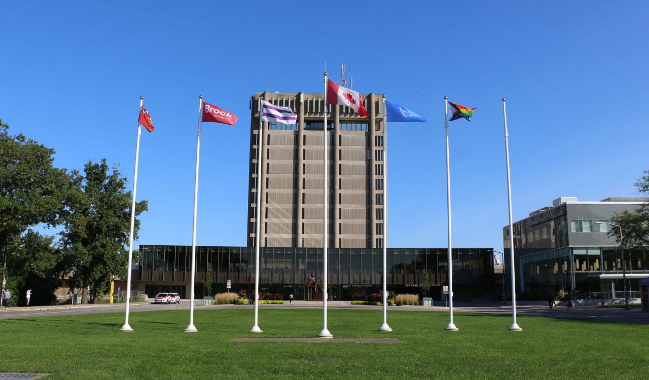 A row of flags stands on an open stretch of grass in front of a large concrete tower.