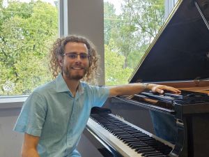 Performance pianist Ryan Baxter sits at a black grand piano in a room with large windows showing green trees outside. Ryan is wearing a light blue button-down shirt and glasses and is smiling warmly at the camera as his arm rests on the piano.