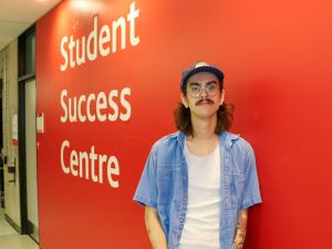 A young man stands in front of a red wall with the words "Student Success Centre" painted on it.