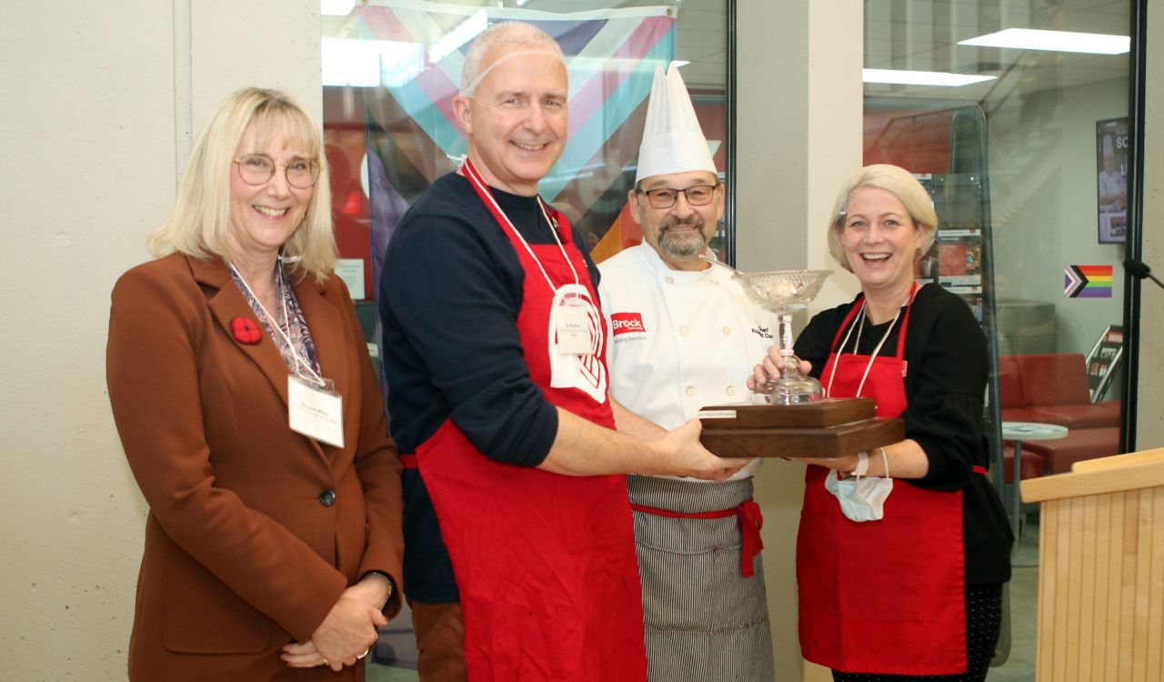 Four people stand next to each other holding a trophy that looks like a clear stemmed bowl with a ladle. One is wearing a chef's hat and whites, and two are wearing red aprons with the United Way logo on them.