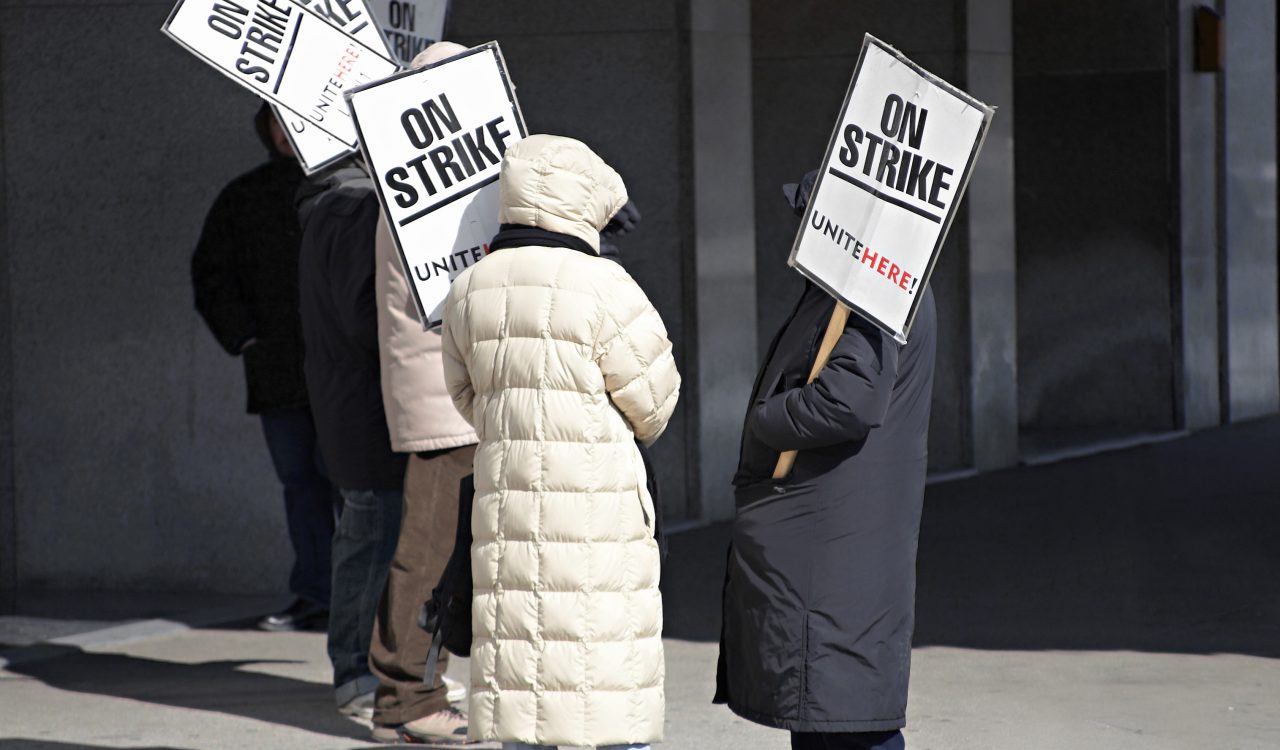 Workers stand outside holding picket signs next to a building.