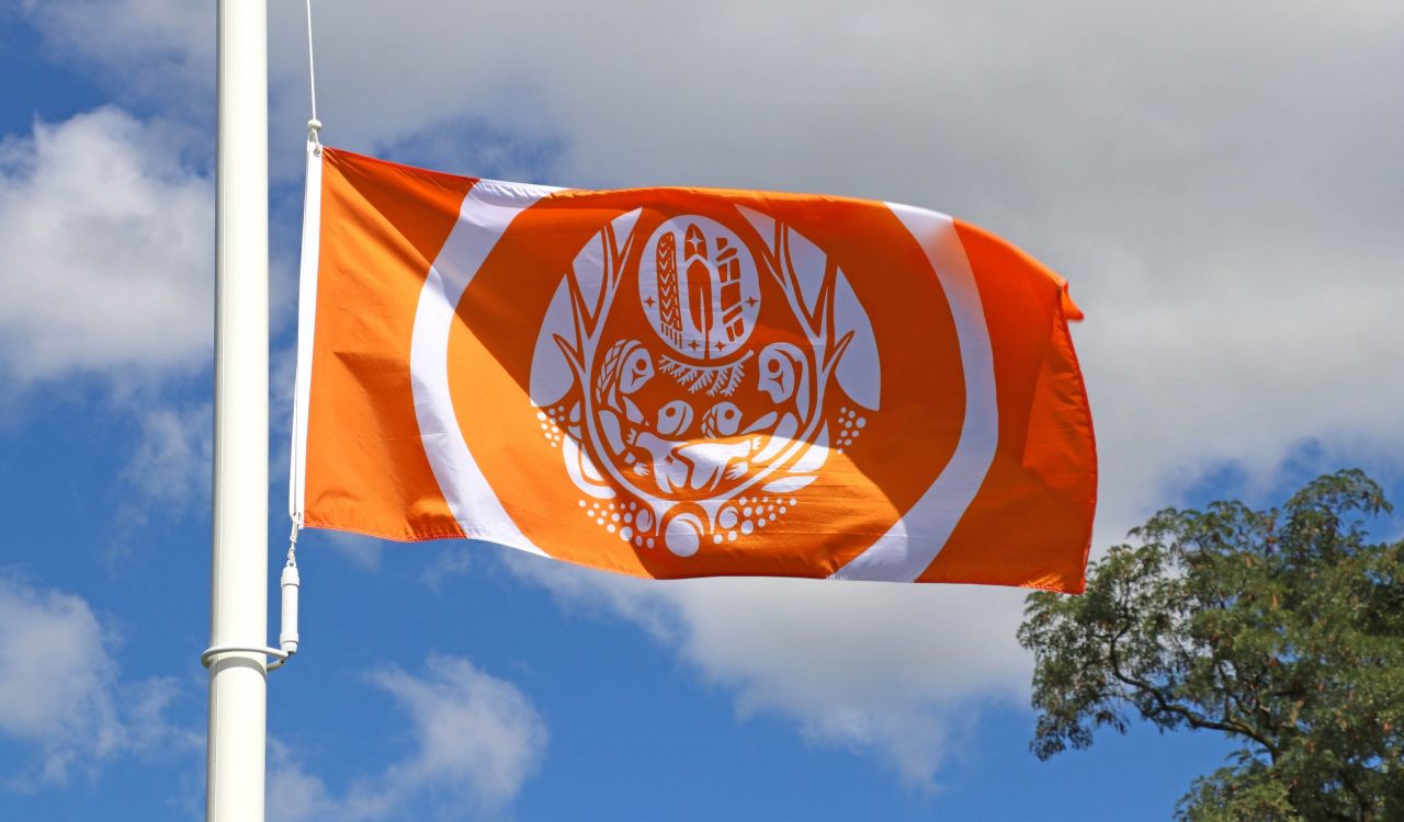 An orange and white flag flies in the wind on a flagpole with trees in the background.