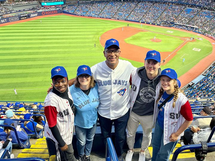 Five people pose for a photo at a baseball game wearing Toronto Blue Jays hats.