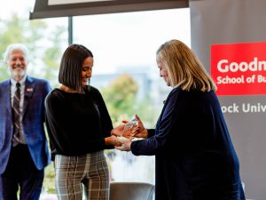 A young student presents a crystal award to another woman on stage in front of a Goodman School of Business at Brock University banner.