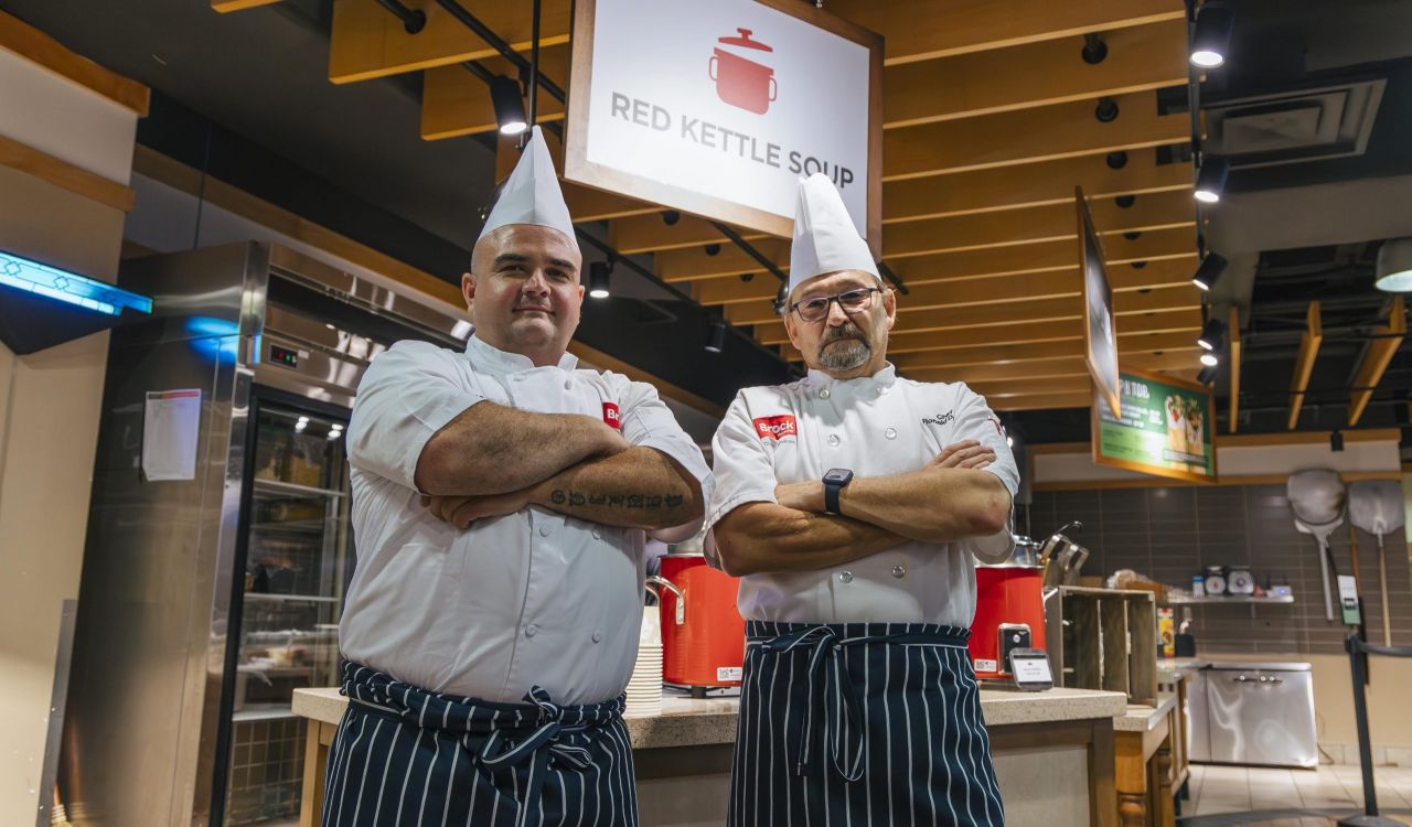 Two men in chef's uniforms stand in a cafeteria with their arms crossed.