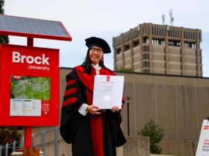 A woman in red and black academic regalia poses for a photo with her diploma outside a university graduation ceremony.