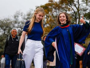 A smiling young woman in a blue academic gown gives a thumbs up while walking with a loved one outside a university graduation ceremony.