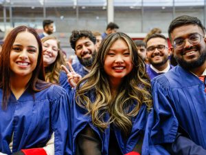 A group of students wearing blue academic robes smile for a photo while sitting in a university graduation ceremony in an auditorium.