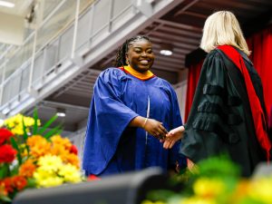 A young woman in a blue academic gown shakes hands with a woman in red and black academic regalia on stage during a university graduation ceremony.