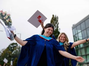 Two young women in blue academic robes smile and cheer outside a university graduation ceremony.