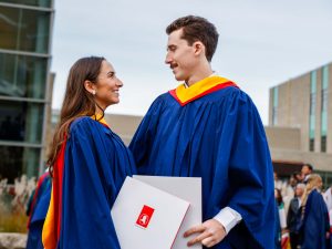 A young man and young woman wearing blue academic robes smile at each other while holding their diplomas outside a university graduation ceremony.