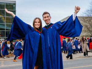 A young man and young woman in blue academic regalia pose smiling for a photo outside a university graduation ceremony.