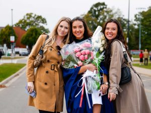 A young woman in a blue academic gown holds bouquets of flowers while posing for a photo with two friends.
