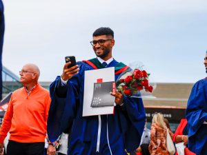 A young man in a blue academic robe smiles at his phone while holding his diploma outside a university graduation ceremony.