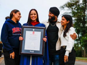 A happy young woman in a blue academic robe poses for a photo with three loved ones while holding a framed diploma outside a university graduation ceremony.