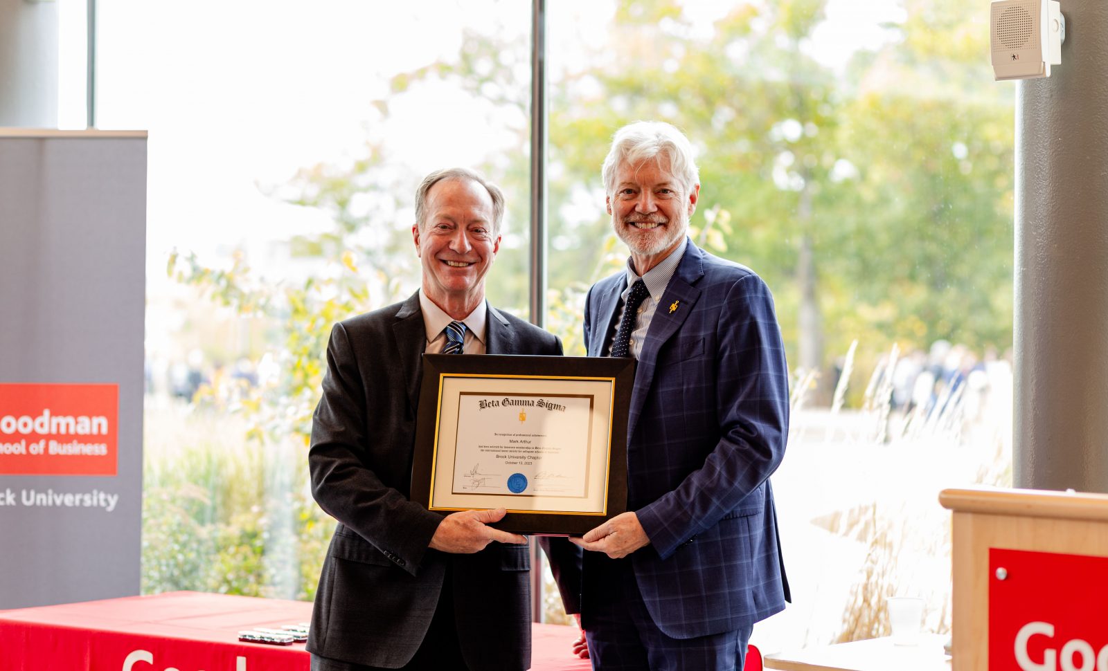 Beside a grey banner and red table cloth that reads Goodman School of Business two men in suits stand together holding a frame with a certificate inside.