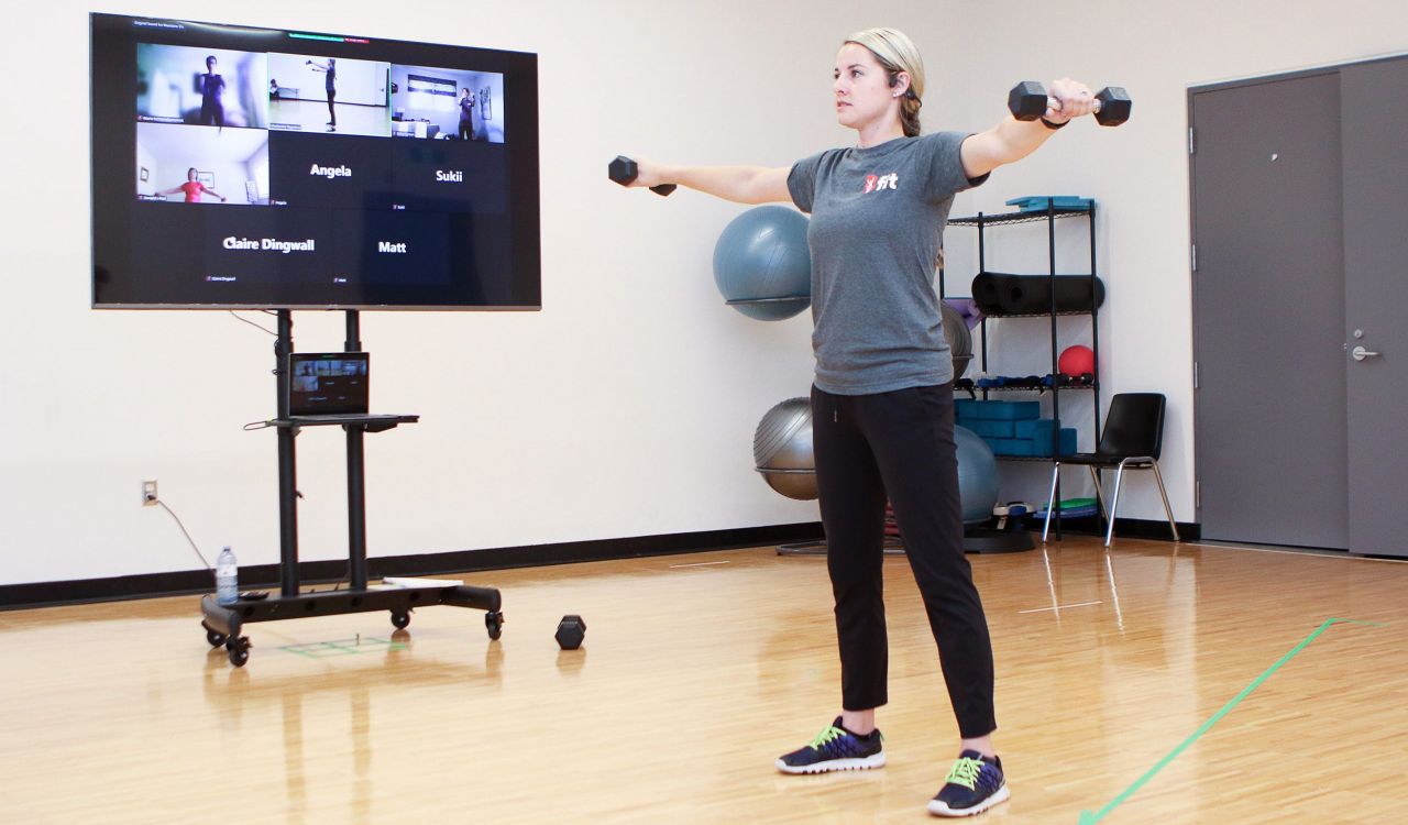 A woman demonstrates exercises for a group of people following along during an online fitness class. Participants can be seen on a television screen.