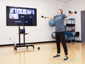 A woman demonstrates exercises for a group of people following along during an online fitness class. Participants can be seen on a television screen.