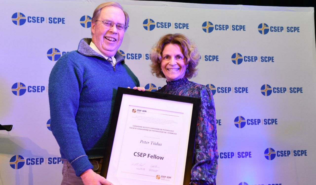 Peter Tiidus and Nota Klentrou hold a large framed “CSEP Fellow” award in front of a photo backdrop filled with a repeating Canadian Society for Exercise Physiology logo
