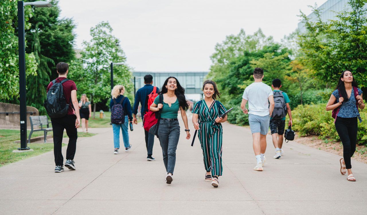 Two women walk together on an outdoor concourse between buildings with trees in the background.