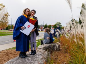Two women stand together on a rock outdoors, one of them in a Convocation gown.
