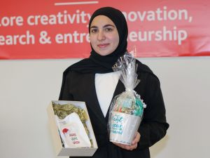 Woman wearing a black headscarf and jacket stands in front of a red sign. She is holding packaged gift in each hand.