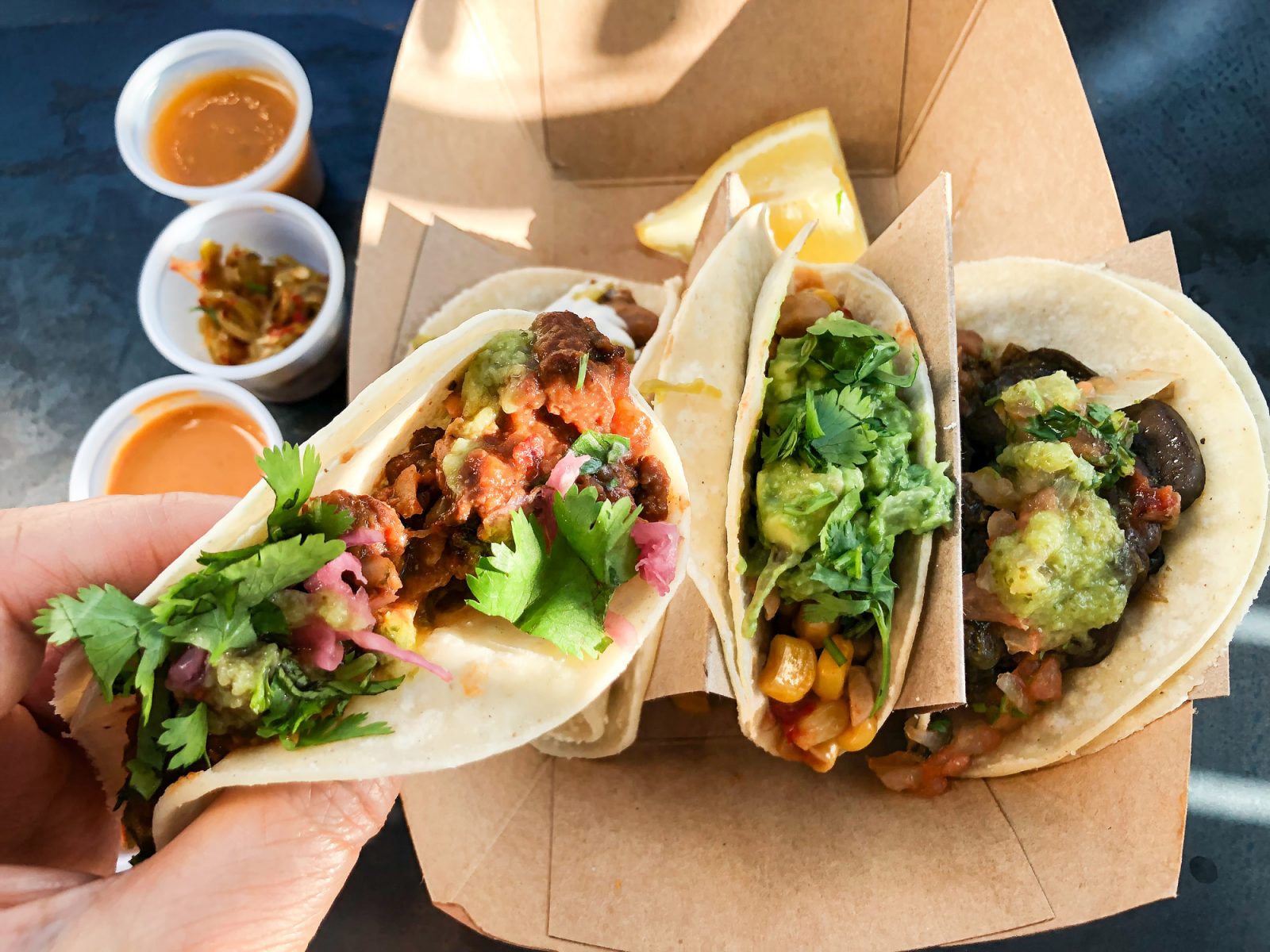A close-up of a hand holding a vegan taco. Two other tacos sit in a take-out container on a table in the background.