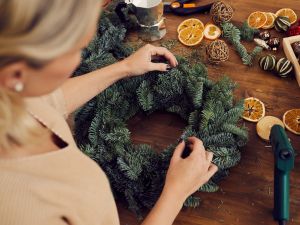 Close-up of unrecognizable woman standing at wooden table and using secateurs while cutting a fir twig for decorative evergreen wreath