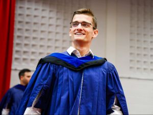 A young man wearing glasses and a blue academic gown smiles as he walks off the stage during a university graduation ceremony.
