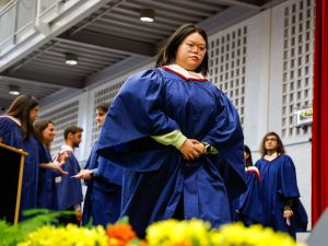 A young woman in a blue academic gown walks across the stage during a university graduation ceremony.