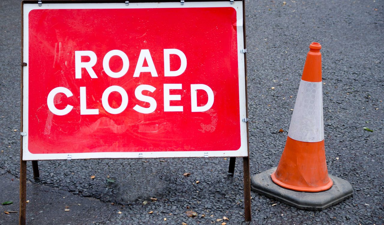 A sign saying ‘Road Closed in white letters on a red background next to an orange traffic cone.
