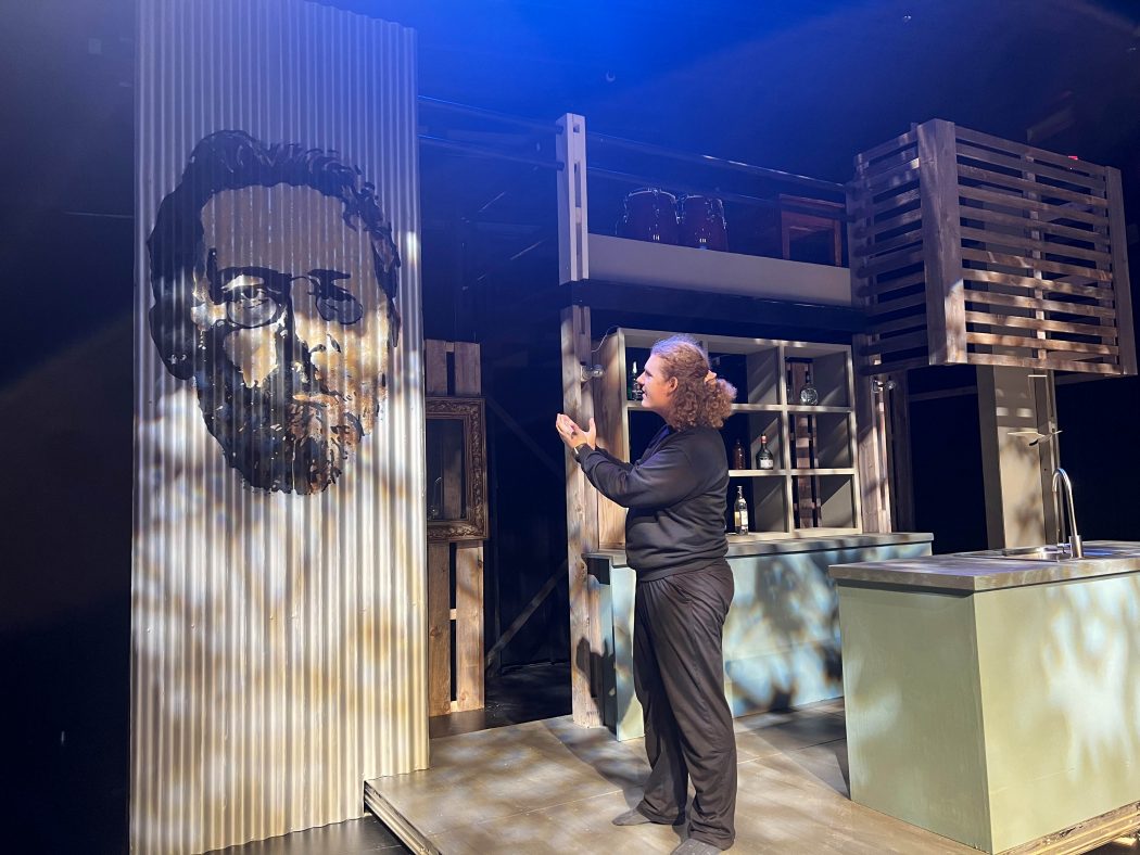 A white man with curly hair dressed in black stands in socked feet on a stage set lit with blue light, with his hands raised as he looks to a stencil on the wall showing an artistic rendering of famed playwright Anton Chekhov.