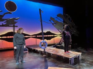 A white man and a black woman stand on a stage set against cool blue light and interesting sculpture made to look like steel in the form of birds. The woman stands on a dock, looking toward the man with a smile. A post with a white lifesaver stands between them.