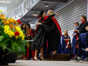 Two women in academic gowns hug on stage during a university graduation ceremony.