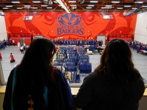 Two people, seen from behind, look down on a gymnasium with racks of academic gowns and students lining up before a university graduation ceremony.