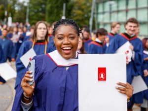 A happy young woman in a blue graduation gown holds up a folder with her university diploma. A crowd of other new graduates can be seen in the background.